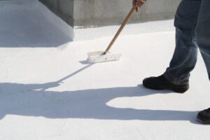A roofing technician applies a white acrylic coating to a flat roof.