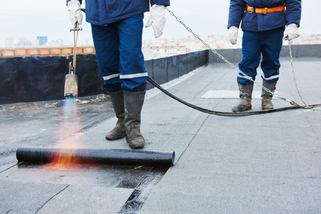 Two roofers work on a flat roof installation by heating melting bitumen roofing felt.