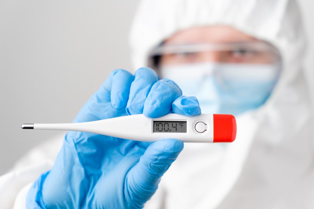 A nurse in protective equipment holding up a thermometer that reads 100.4 degrees.