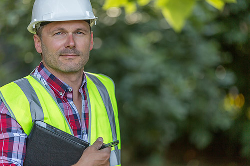 A roofer carries a tablet in a reflective vest and hardhat.