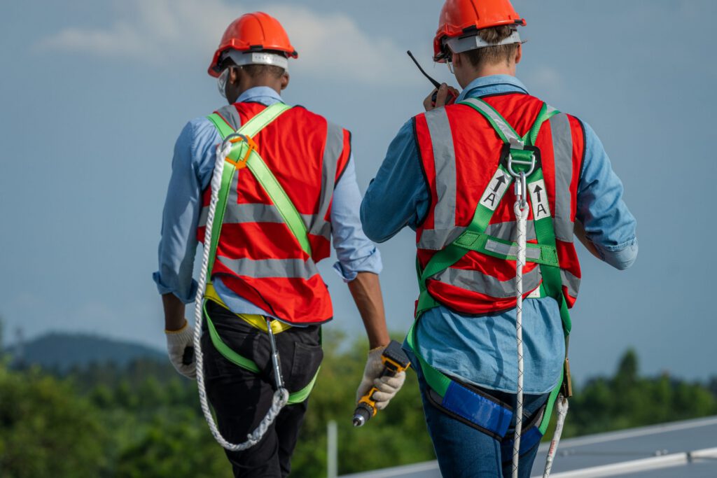 A pair of roofers in full PPE and safety equipment walk away from the camera, as one speaks into a radio