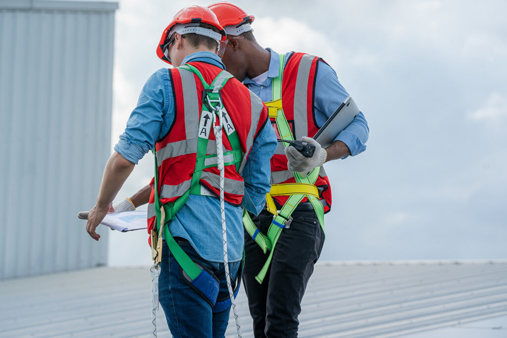 Two men in safety harnesses and vests discuss their commercial roof inspection checklist with their backs to the camera