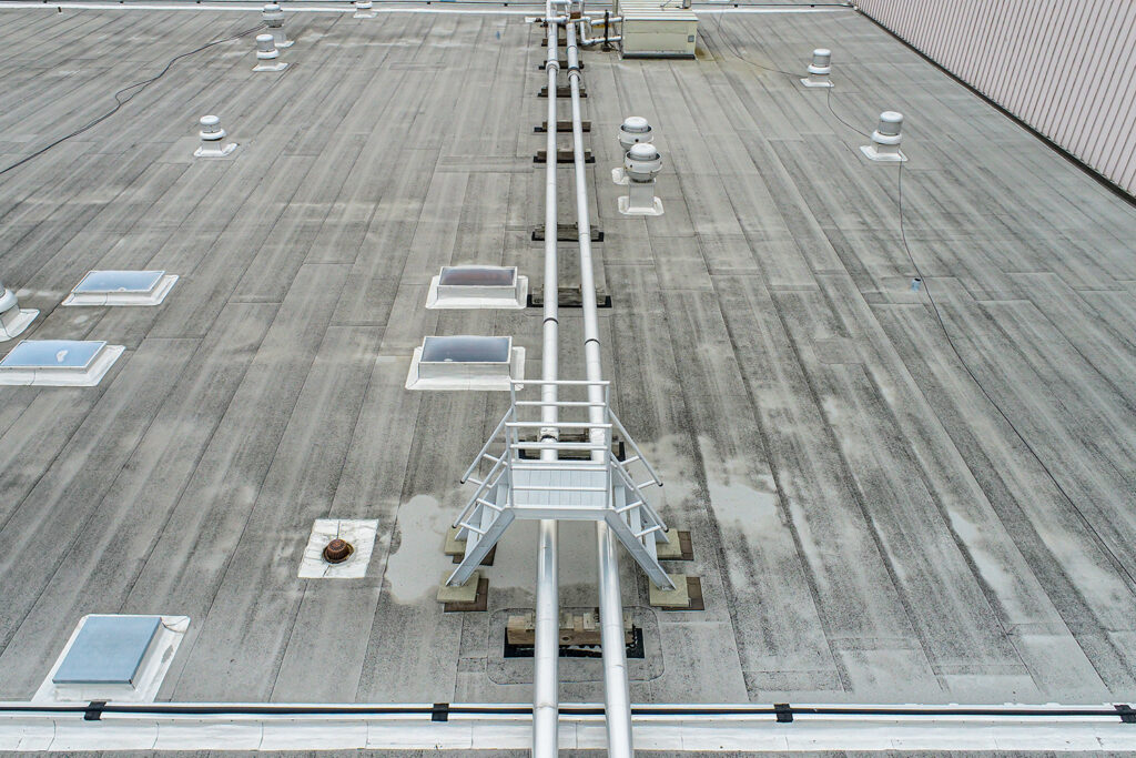 A view from above of a gray commercial roof bisected by two pipes running along the center with a short pair of stairs leading over them.